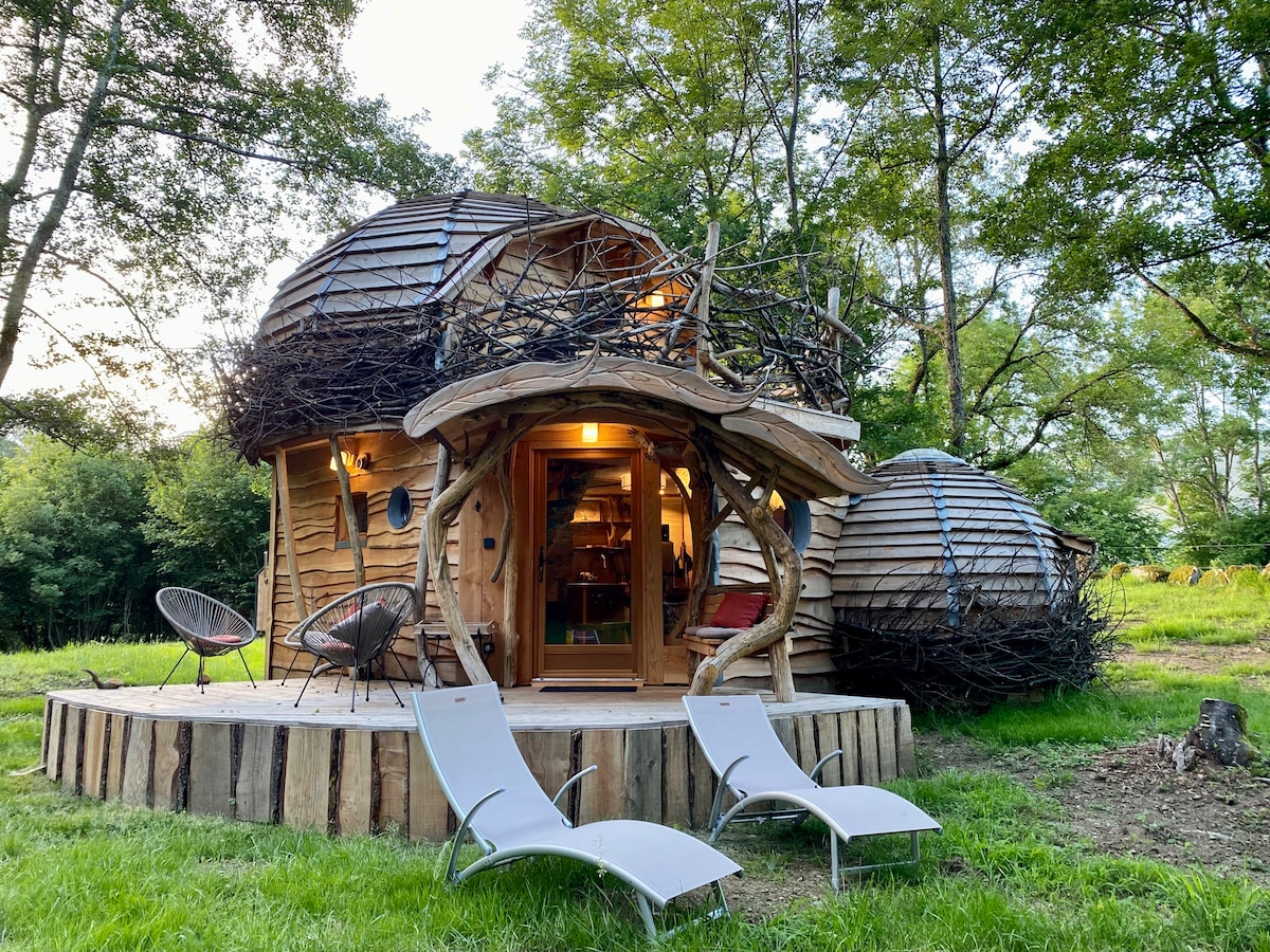 An original wooden cabin is surrounded by greenery, featuring a unique architectural design with natural materials. Two lounge chairs are positioned in the foreground, inviting relaxation. The cabin is accentuated by a rounded shape and is complemented by a welcoming entrance.