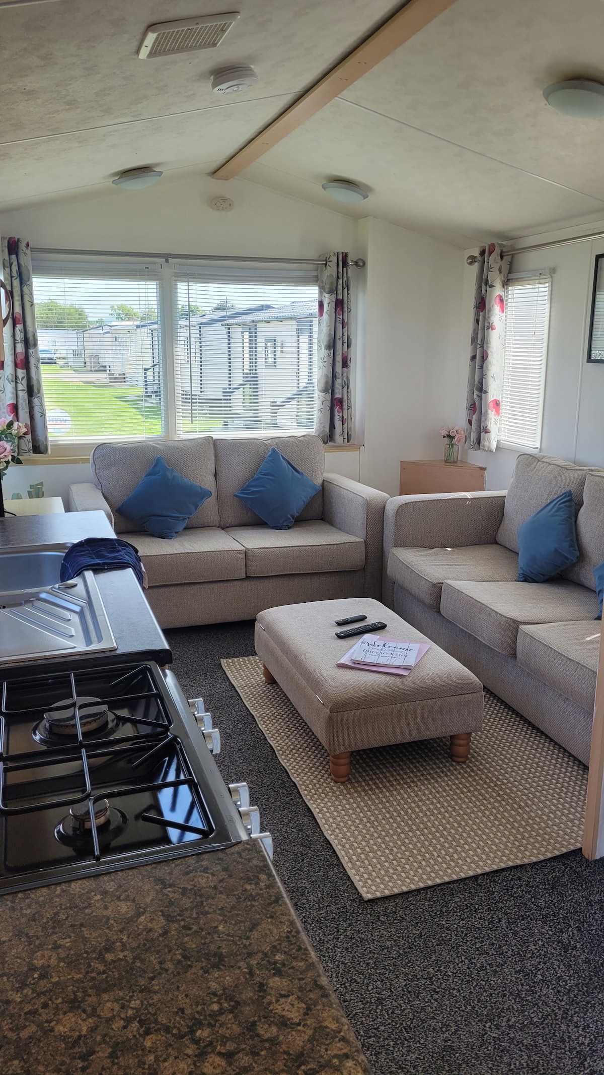 The living area features a cozy sofa with two blue throw pillows, complemented by a matching footrest. Large windows allow natural light to fill the space, while a small table holds a magazine. A stove is visible in the kitchen area adjacent to the seating.