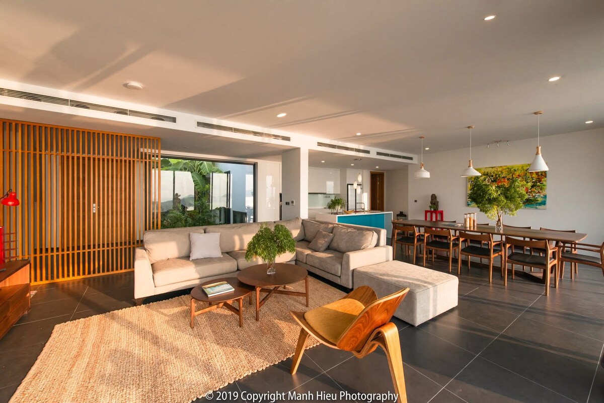 An open-concept living area features a light-colored sofa and a wooden coffee table on a textured rug. A dining table is visible in the background. Large windows provide views of the surrounding landscape, while natural light fills the space, emphasizing the modern design.