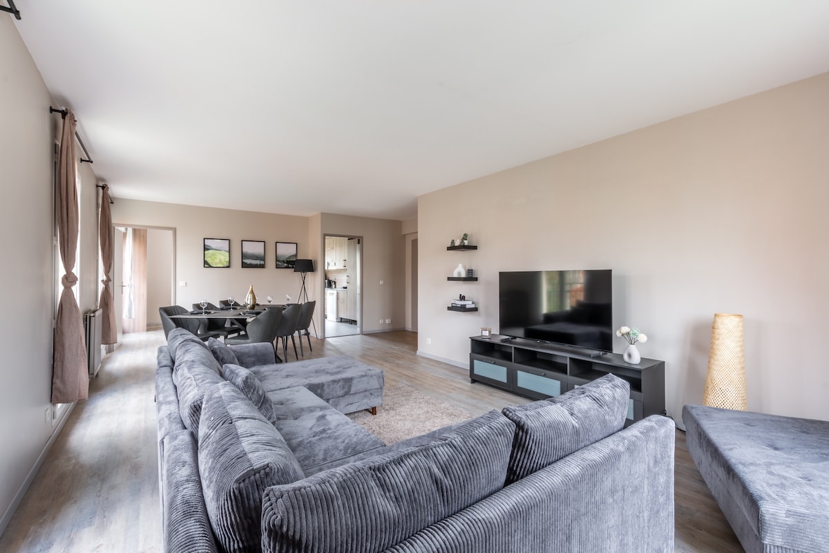 A spacious living area is furnished with a large sectional sofa and a cozy armchair. A flat-screen television is positioned against the wall, accompanied by minimalistic shelving. A dining area for six is visible in the background, next to an open kitchen that offers easy accessibility.
