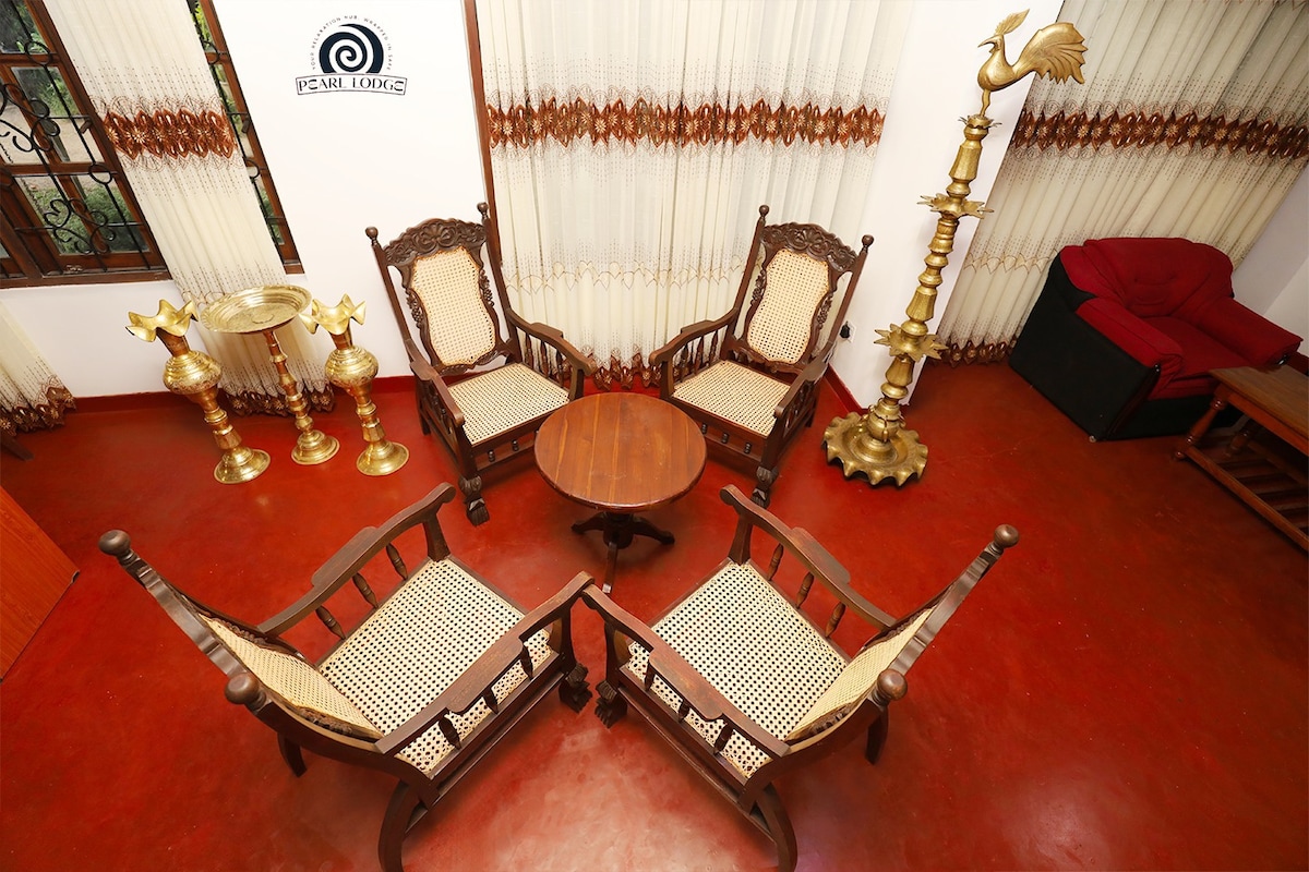An elegantly furnished seating area is shown, featuring four carved wooden chairs with intricate designs and traditional woven seats arranged around a round wood table. Soft lighting is provided by two decorative brass lamps, and large curtains frame the windows, creating a comfortable atmosphere.