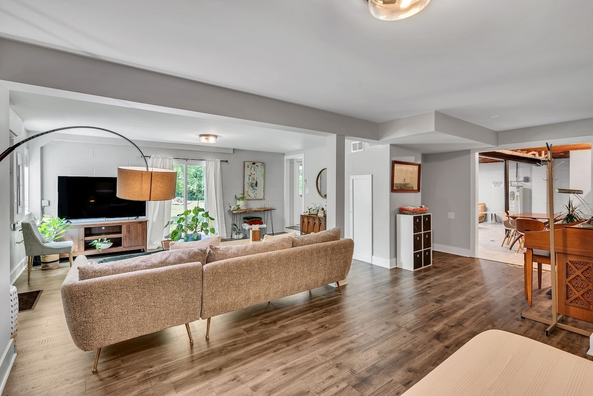 An open living space is featured with a large sectional couch and a mounted television. Natural light is welcomed through multiple windows, while potted plants add a touch of greenery. A dining area with wooden furnishings is visible in the background, enhancing the communal atmosphere.