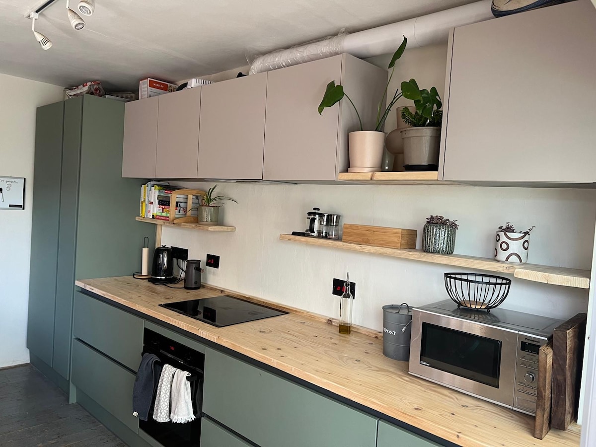 A modern kitchen is displayed, featuring sleek cabinetry in muted green and light grey. Wooden countertops provide space for various appliances, including a microwave and a kettle. Shelves lined with potted plants and decorative items add a touch of warmth and practicality.