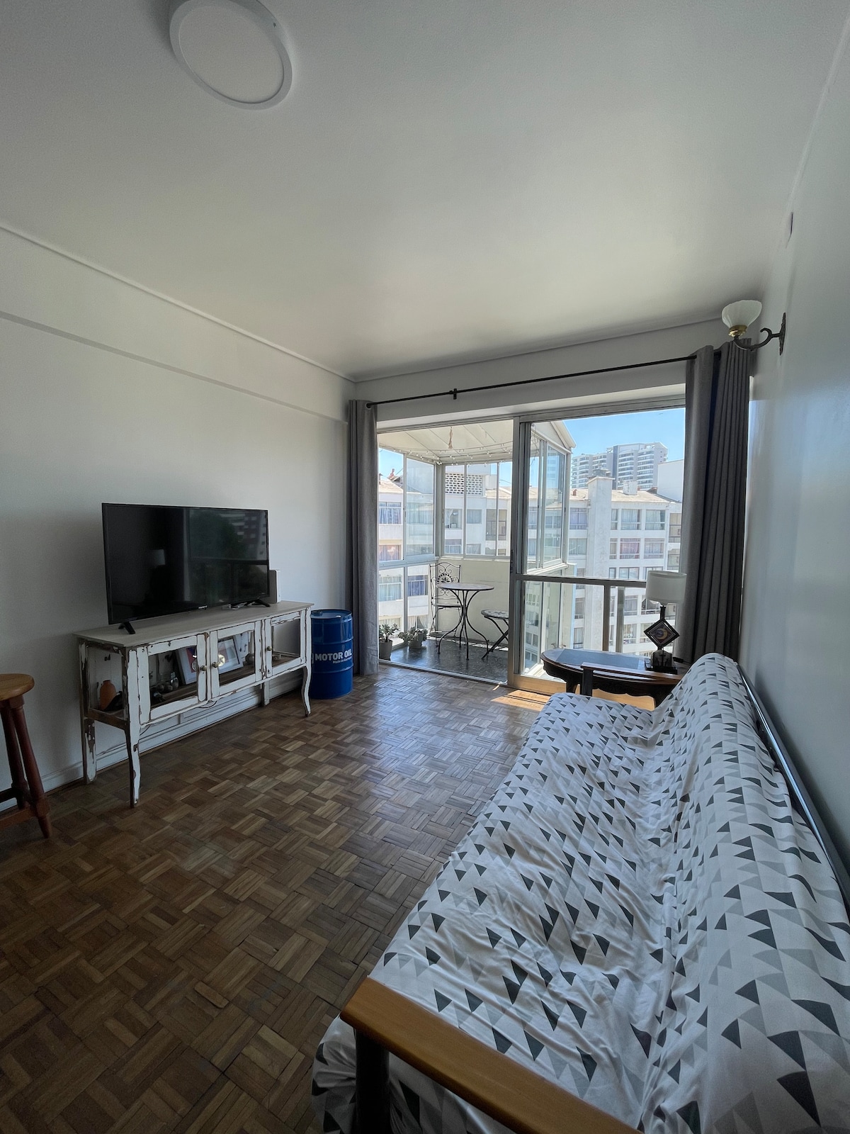 A light-filled living area features a patterned sofa facing a television placed on a white console. A glass door leads to a small balcony with outdoor seating, while large curtains frame the window, allowing natural light to brighten the space.