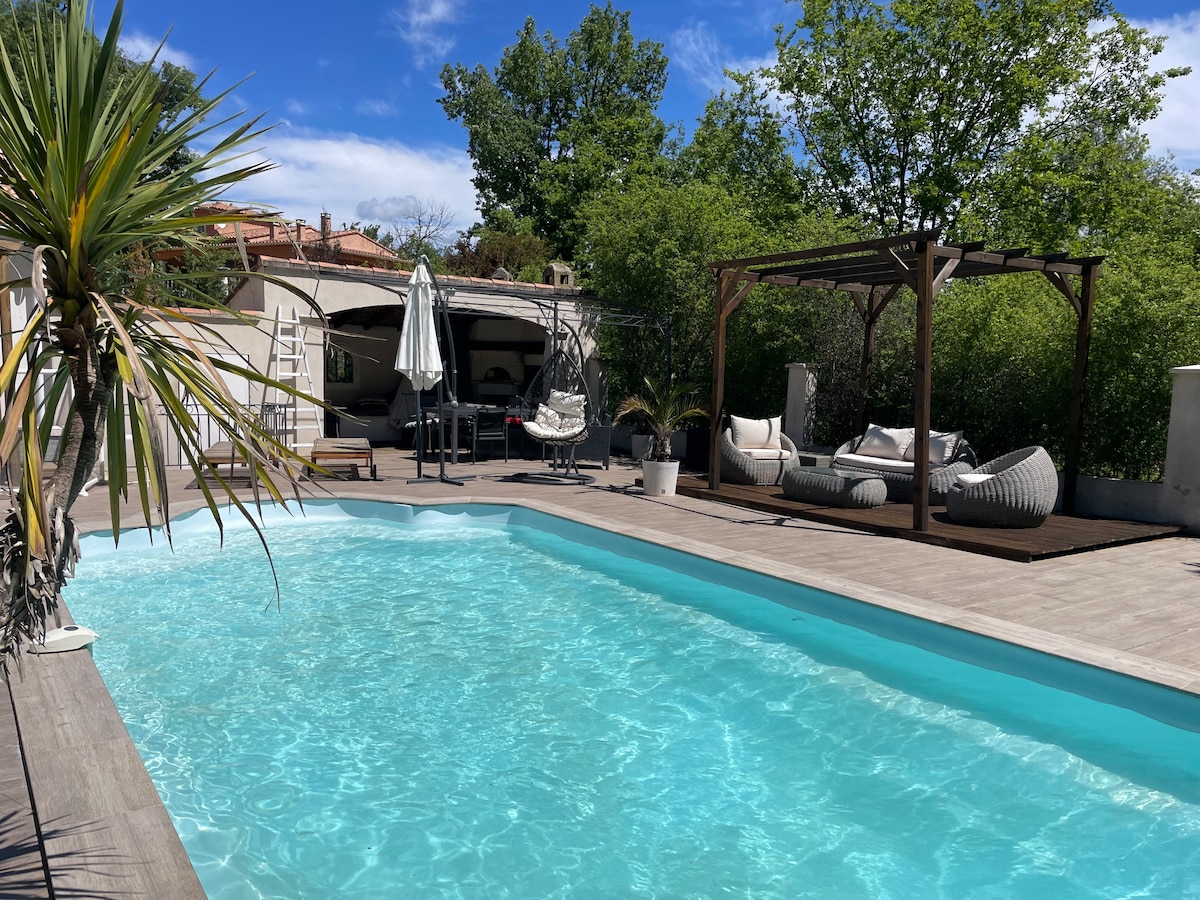 A private swimming pool is surrounded by greenery, featuring a light blue water surface. Lounging areas with comfortable seating, including a shaded pergola with swinging seats, are visible. Sun loungers and an umbrella provide additional relaxation options by the poolside.