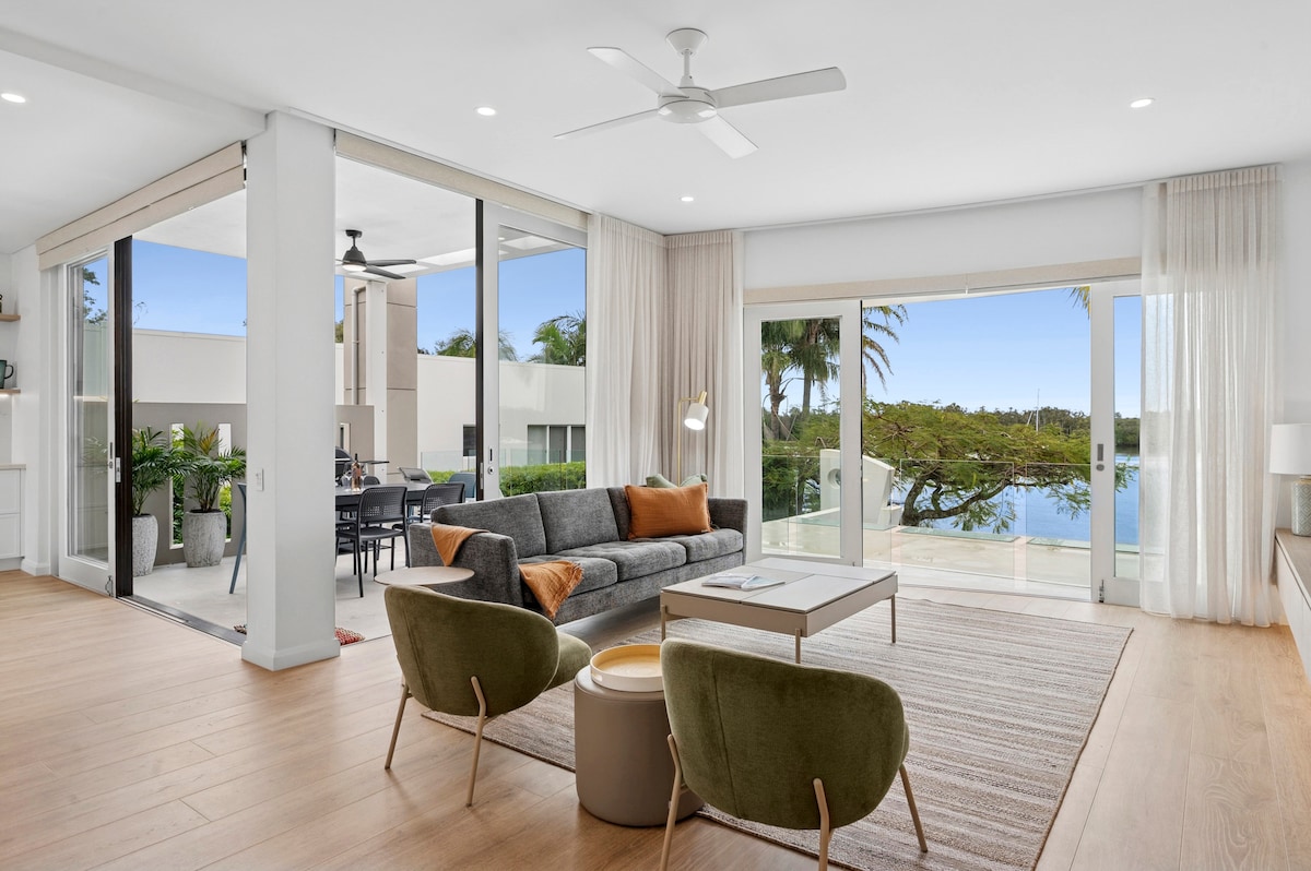 A spacious living area features large glass doors that connect indoor and outdoor spaces. A comfortable gray sofa and two green accent chairs are arranged on a light wood floor, complemented by a neutral area rug. Natural light floods the space, showcasing views of palm trees.