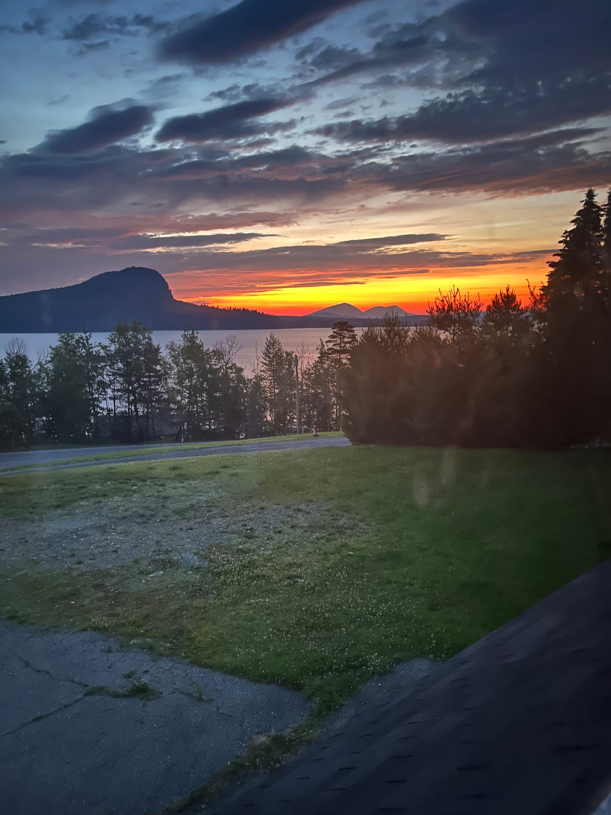 A scenic sunset view is captured, showcasing vibrant colors reflecting off the lake. Majestic Mt. Kineo is visible in the background, framed by greenery. The foreground features a grassy area with a clear path leading to the water, enhancing the natural beauty of the landscape.