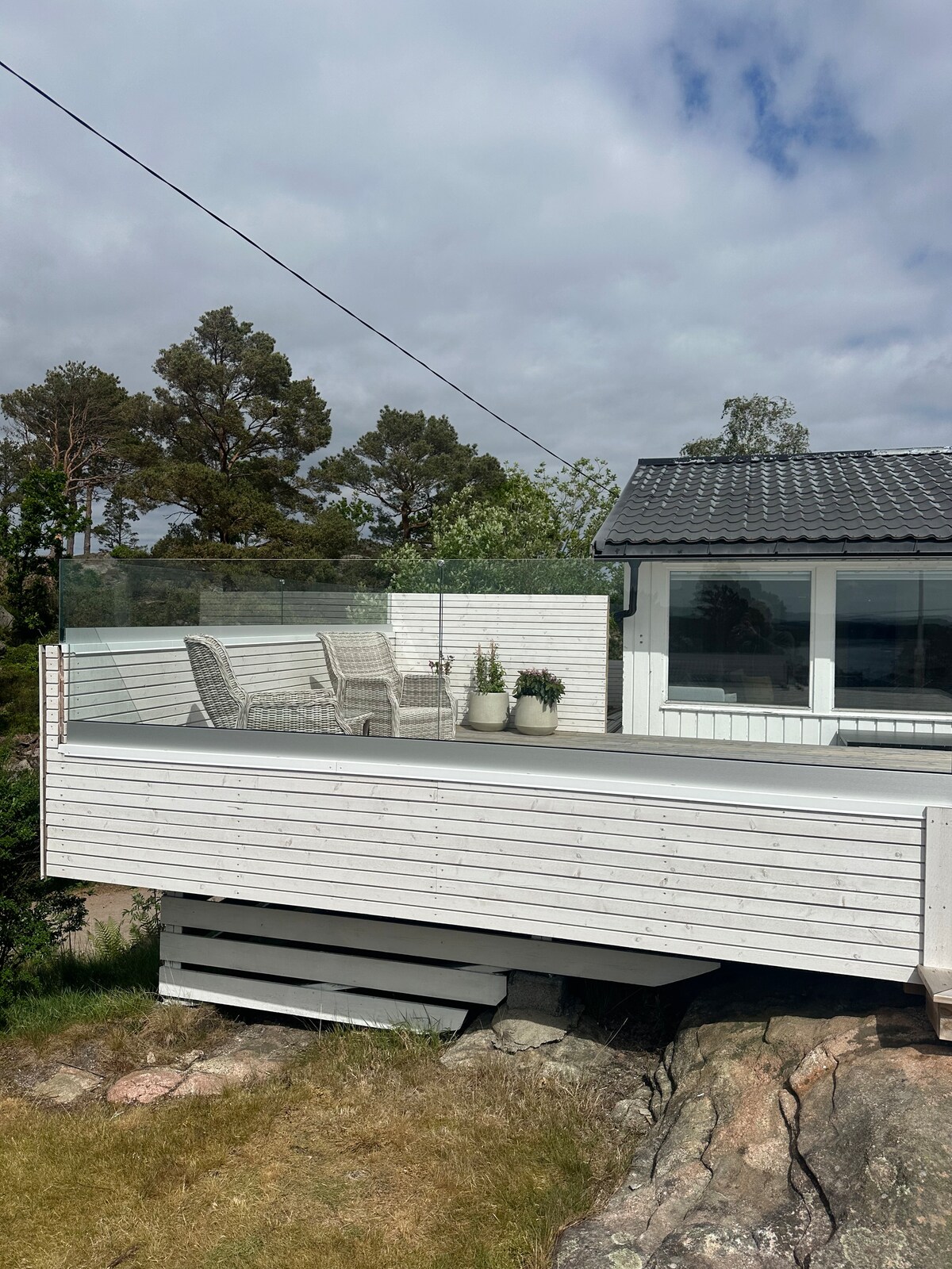 A spacious terrace is shown, featuring a glass railing that offers unobstructed views. Two wicker chairs are positioned on the deck, surrounded by potted plants. The exterior of the cabin exhibits a light-colored wood finish, blending harmoniously with the natural surroundings.