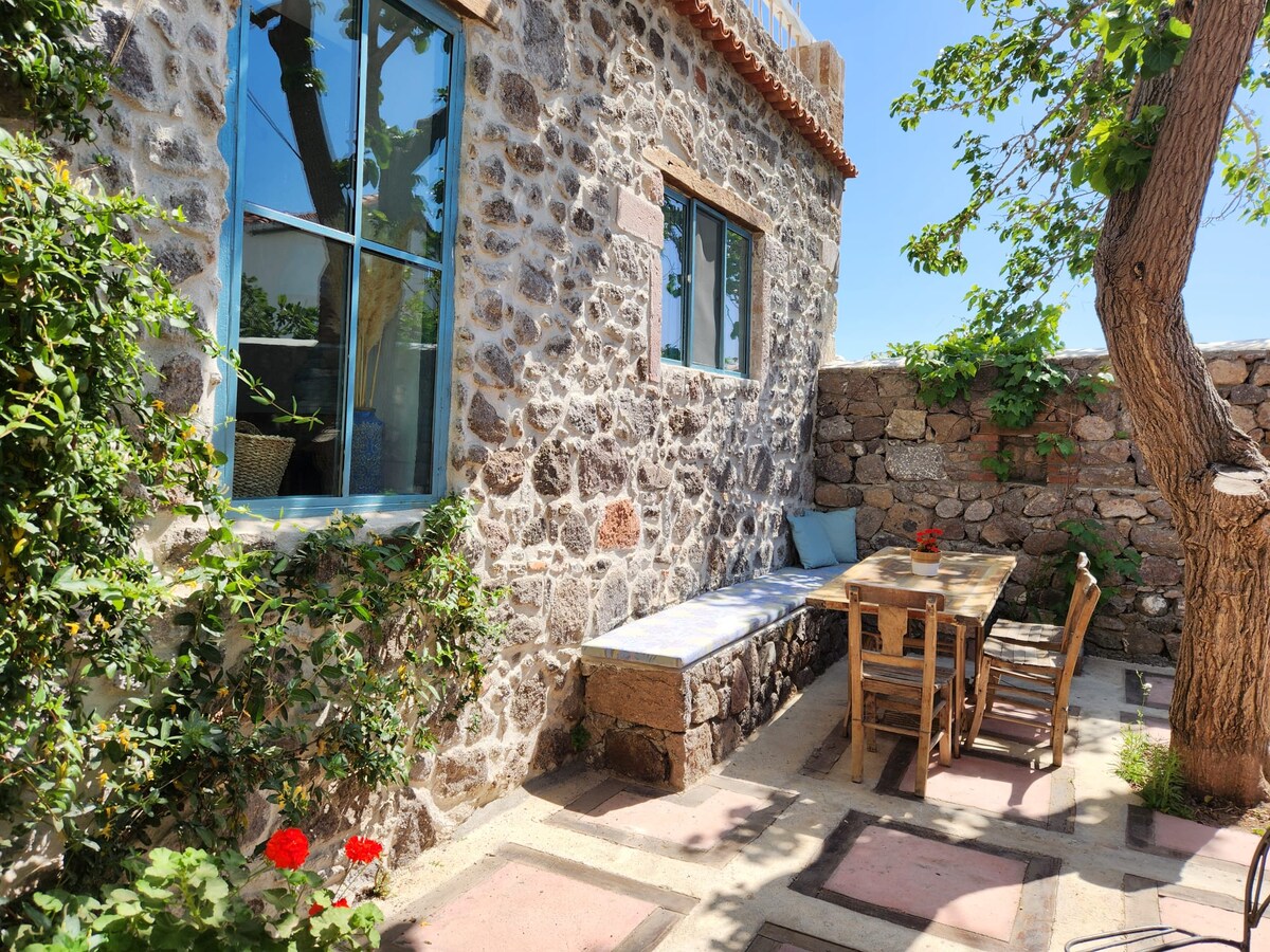 An outdoor dining area is featured, surrounded by stone walls and greenery. A wooden table is set with several chairs, and a cushioned bench runs along the wall. Sunlight casts gentle shadows, highlighting the rustic character of the space.