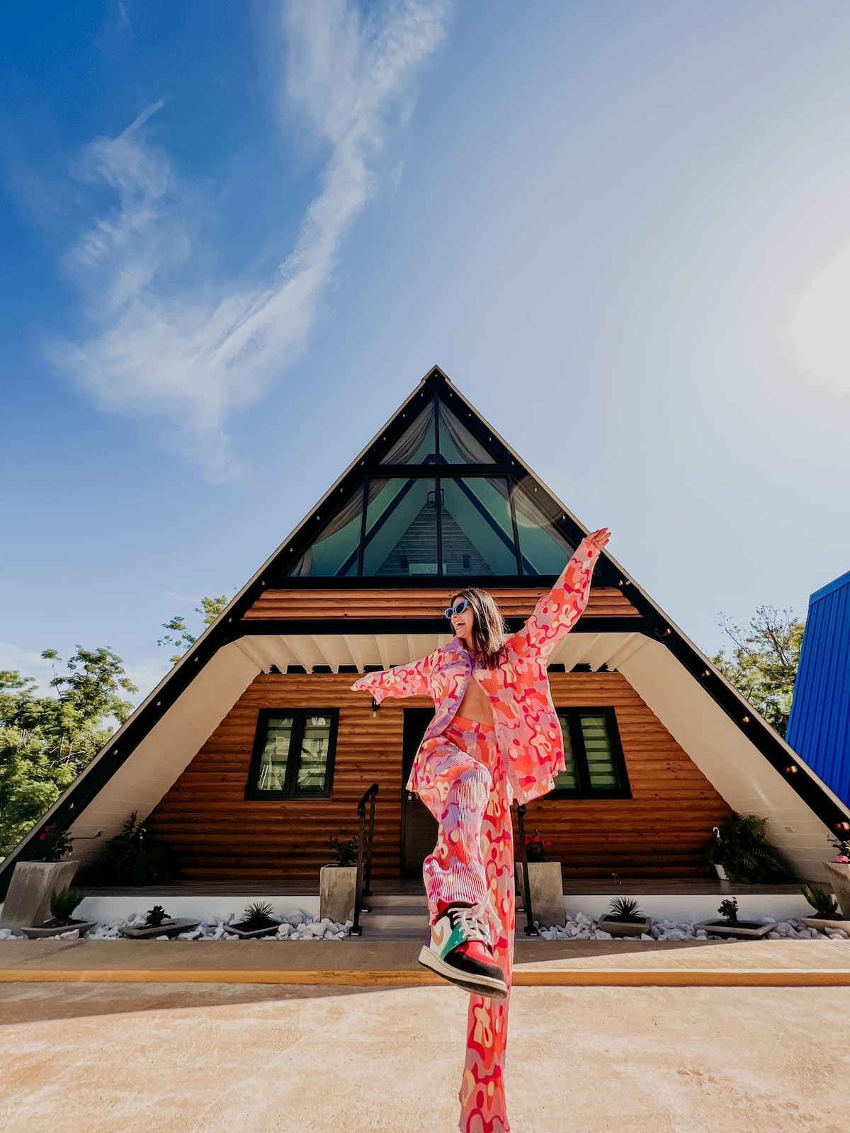 An A-frame villa is showcased with a triangular wooden structure, large glass windows, and a welcoming entrance. Lush greenery surrounds the property, enhancing its natural setting. The space is bathed in sunlight, creating an inviting atmosphere for guests.