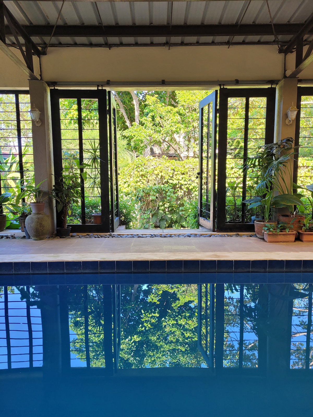 Large glass doors open to reveal a serene view of the lush garden, with greenery reflected in the clear water of the indoor pool. Potted plants line the edges, adding an element of nature to the inviting space.