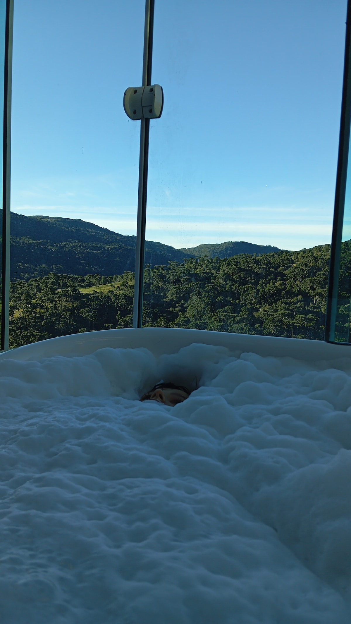 A large freestanding tub is filled with bubbles, positioned in front of floor-to-ceiling windows. The serene view of lush green mountains is visible in the background, complementing the peaceful ambiance of the bathing area.