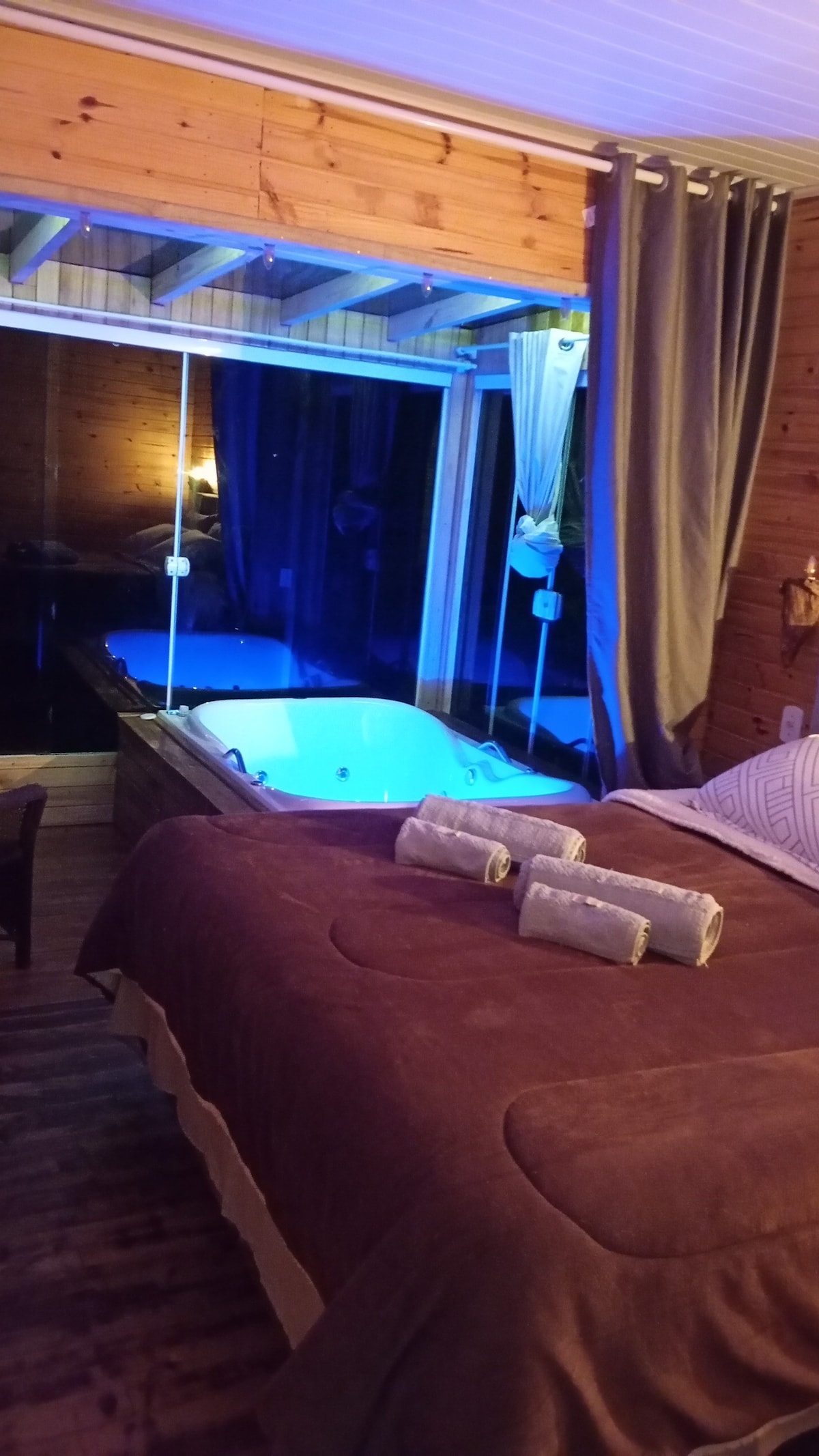 A serene interior features a spacious bed adorned with a soft brown blanket. Next to the bed, a bathtub is presented with a soothing blue light. Large glass doors provide a view of the outside, while curtains frame the window, adding a touch of privacy.