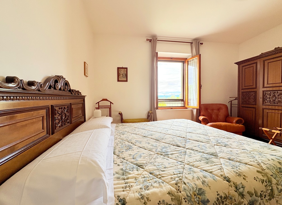 A spacious room features a large bed with a floral-patterned coverlet, complemented by a wooden headboard. Natural light floods in through an open window, providing a view of the outside. Additional furniture includes a wooden armchair and a tall wardrobe.