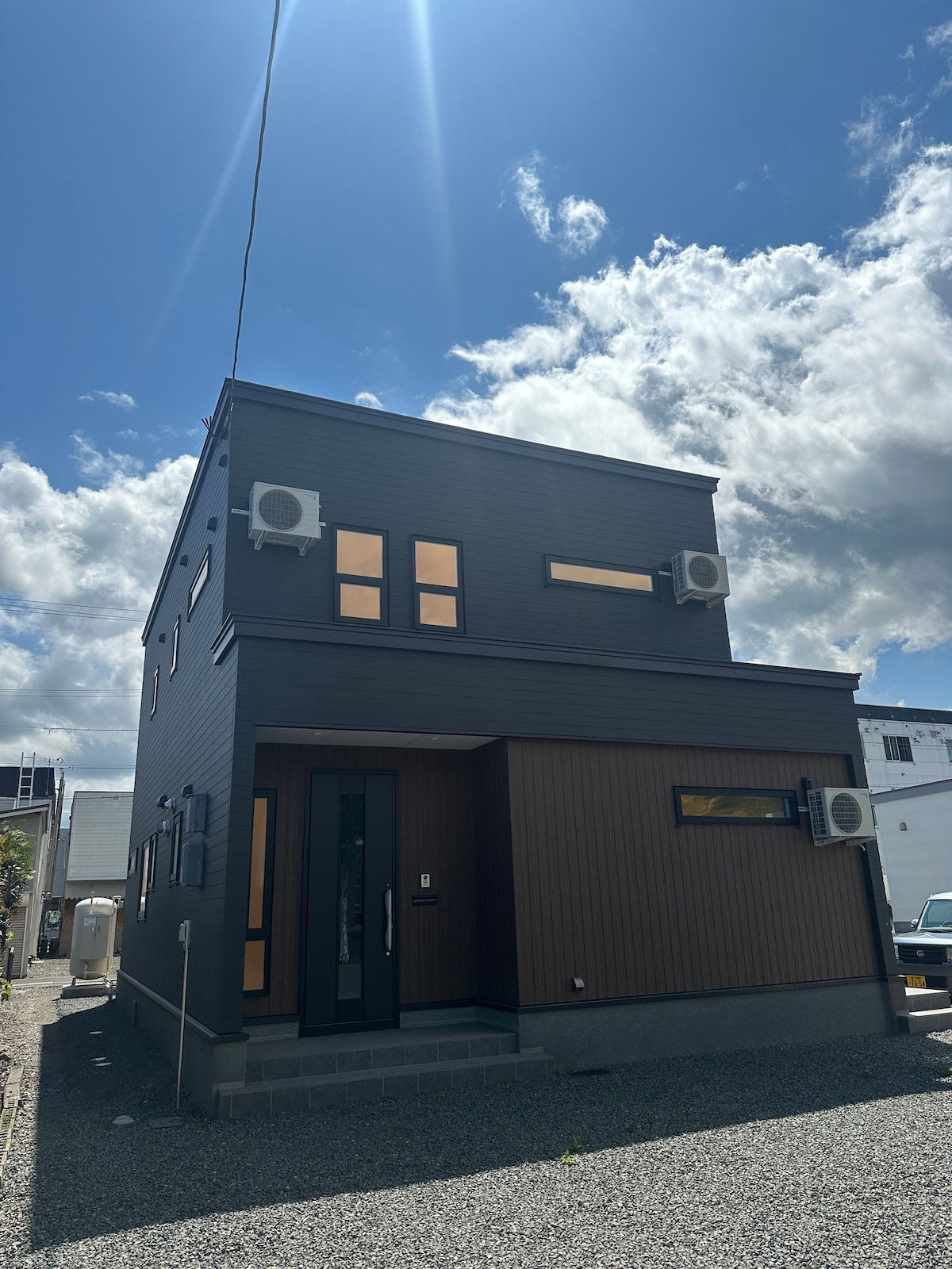 The modern two-story house features a combination of dark wood and sleek black siding. Large windows provide ample natural light, while multiple air conditioning units are visible on the exterior. The property is surrounded by a gravel pathway, enhancing the urban setting.