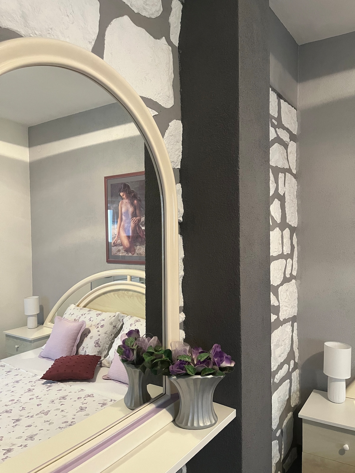A mirror reflects a cozy bedroom space with a light-colored bed dressed in soft, patterned linens. A colorful wall art piece is visible alongside a vase of purple flowers on a bedside table, complemented by modern lamps on either side.