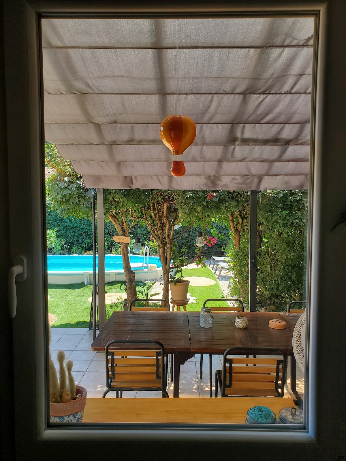 A view from a window captures an outdoor dining area beneath a shaded pergola. A wooden table is surrounded by several chairs. Beyond, a blue above-ground pool is visible amidst greenery and ornamental plants, creating a peaceful outdoor setting.