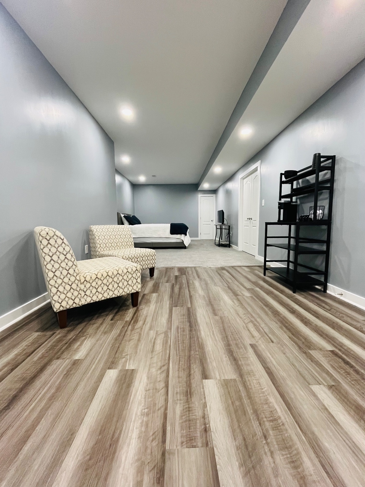 The spacious studio basement features light grey walls and an appealing wooden plank floor. A seating area with patterned chairs is positioned next to a sleek black shelving unit. A large bed is situated in the background, enhancing the open layout.