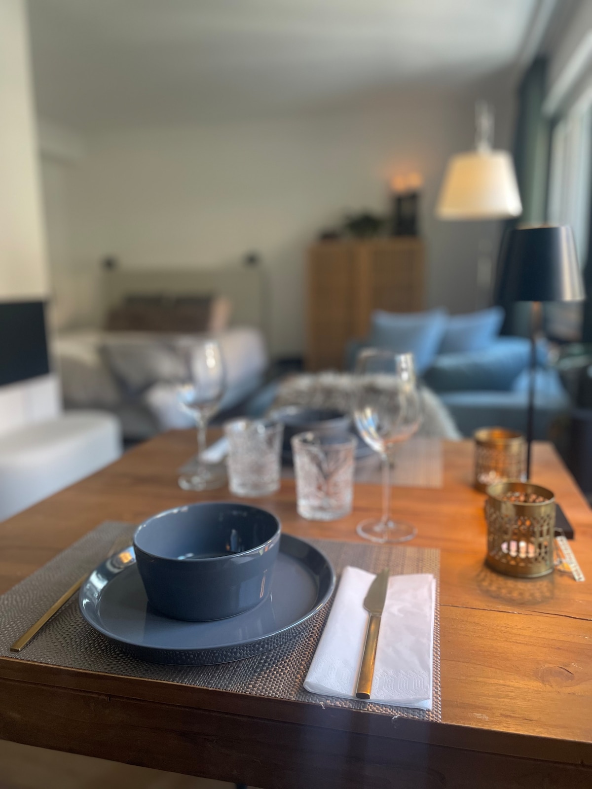A dining table is set with a dark bowl and matching plate, accompanied by elegant glassware. Soft lighting and a cozy setting are provided by a floor lamp nearby. In the background, a spacious bed is visible, complemented by a blue armchair.