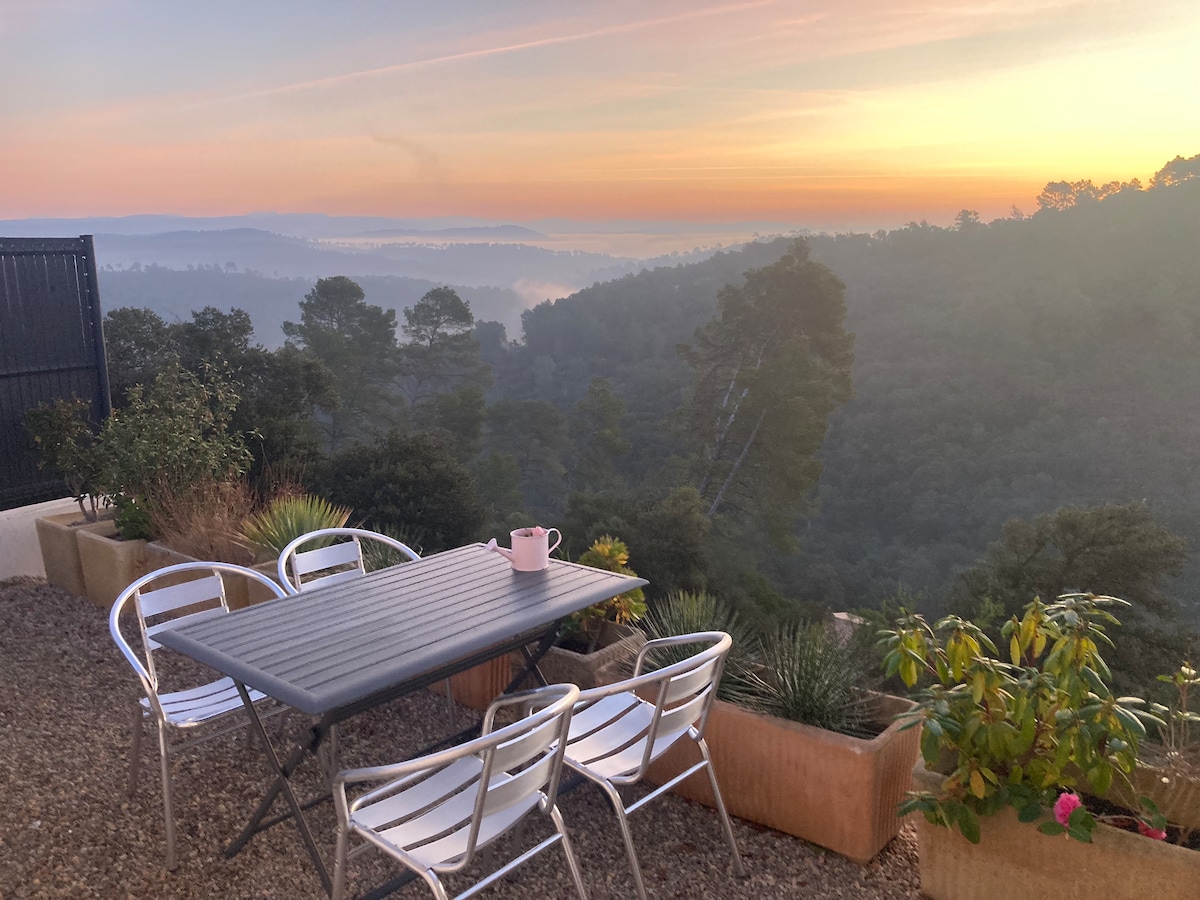 An outdoor terrace features a metal table set for four, surrounded by planters with greenery. The view overlooks rolling hills shrouded in morning mist, with a soft sunrise illuminating the sky in pastel hues.