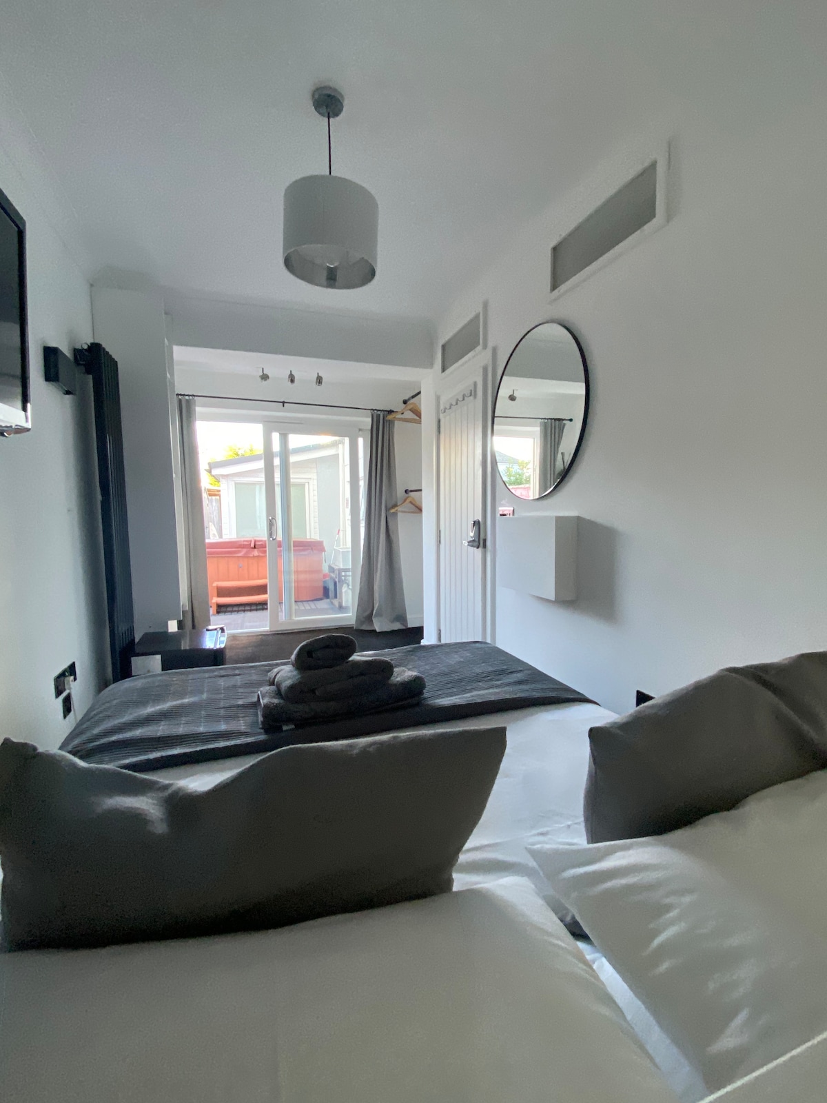 A well-appointed bedroom features a spacious bed adorned with neatly arranged towels. Natural light fills the room through sliding glass doors that open to a outdoor space. A round mirror is mounted on the wall, and a television is positioned nearby.
