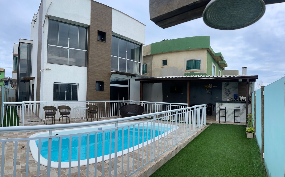 An outdoor area features a fenced pool surrounded by a patio, complemented by lounge chairs and a grassy space. A covered barbecue area is visible, along with large windows of the adjacent building, enhancing the inviting atmosphere for outdoor gatherings.