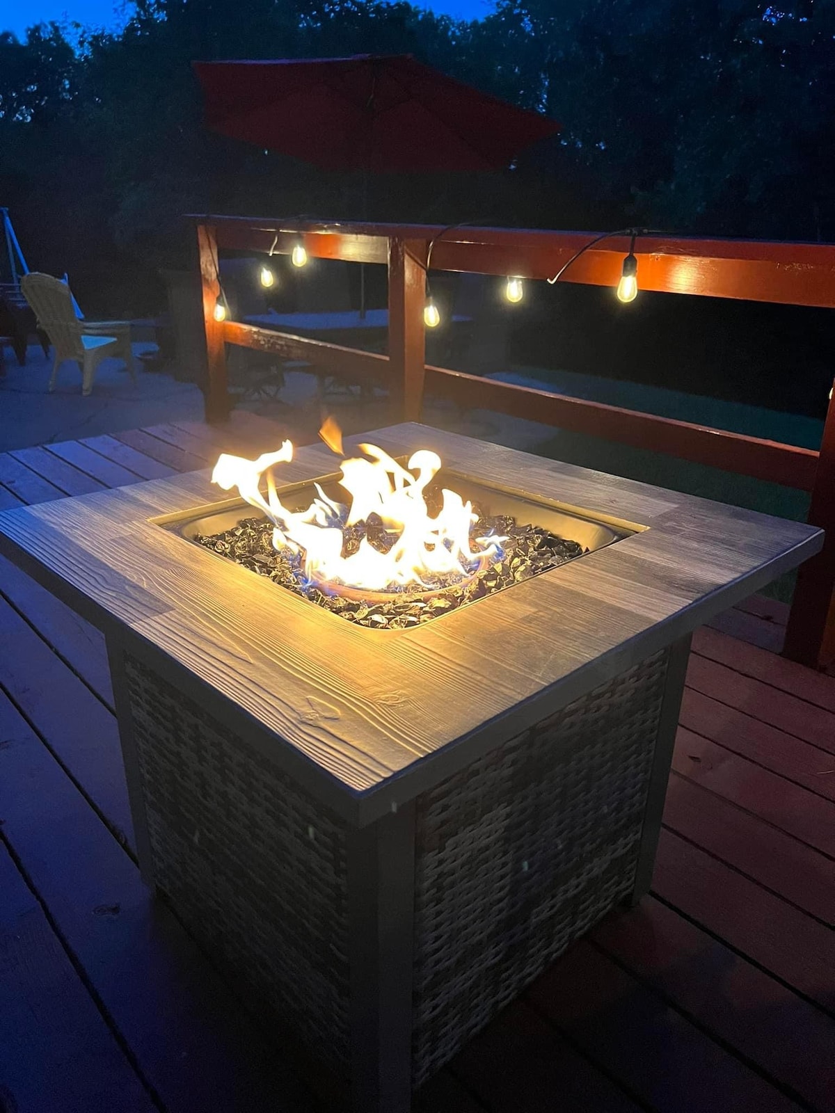 A fire pit is centrally located on a wooden deck, surrounded by a woven base and filled with decorative stones. Soft, twinkling lights hang above, creating a warm ambiance. In the background, lounge chairs are visible, positioned for relaxation.