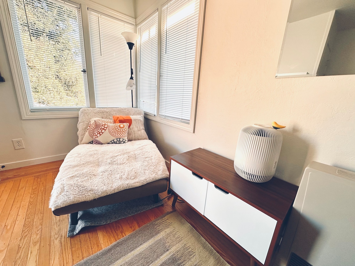A comfortable seating area features a cozy chair adorned with decorative pillows and a soft throw blanket. Natural light fills the space through large windows with blinds. A sleek wooden media console stands nearby, complemented by a modern air purifier.
