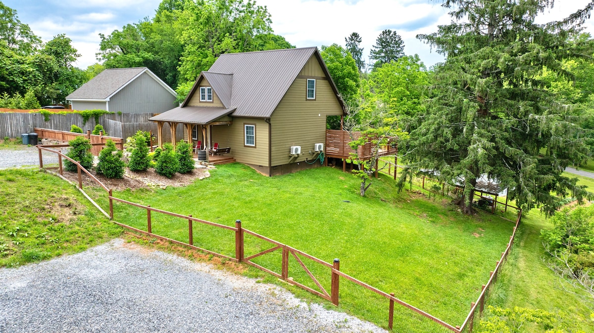 A charming two-story house is surrounded by a spacious, fenced yard featuring well-maintained grass and garden beds. The exterior includes a covered porch and a gravel parking area, with trees providing a natural backdrop to the property.