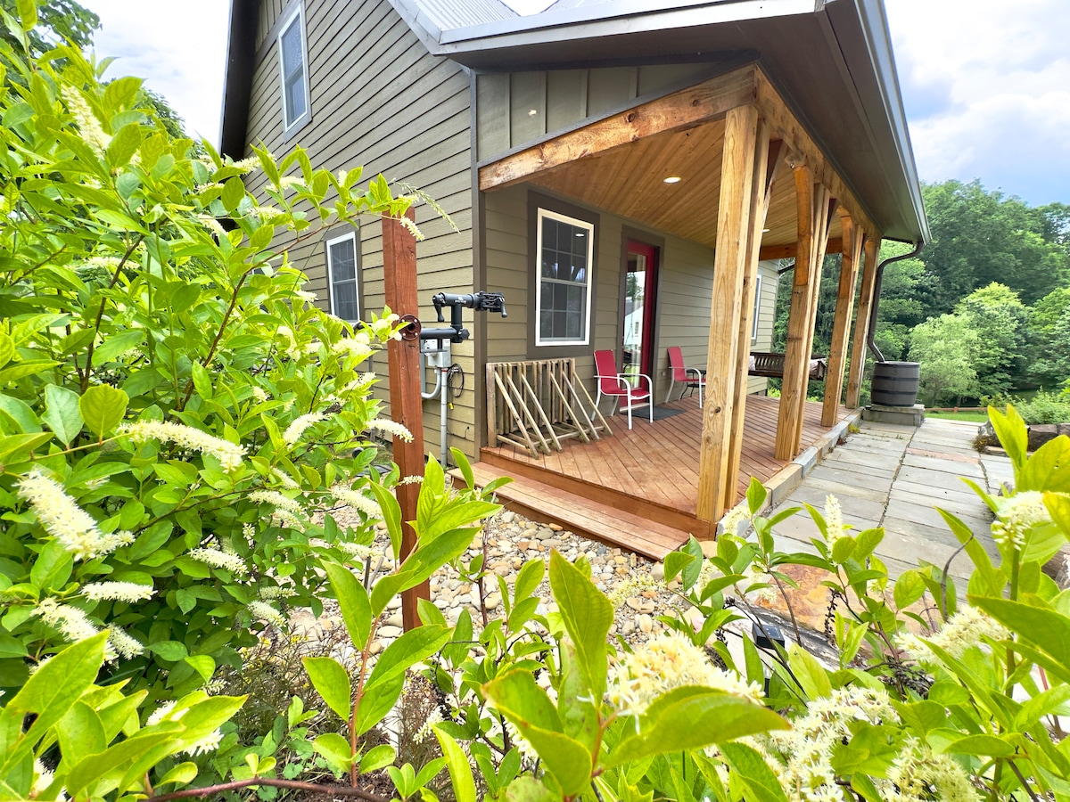 A welcoming porch is featured at the entrance of the house, surrounded by lush greenery. Two chairs are positioned on the wooden deck, providing an inviting space for relaxation. The home’s natural tones blend harmoniously with its outdoor setting.