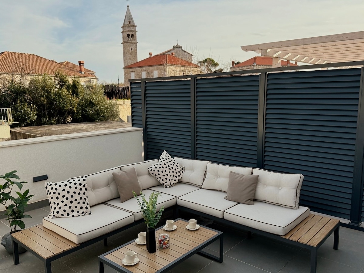 A spacious terrace features a modern seating arrangement with a sectional sofa and several cushions. A wooden coffee table holds a small decorative plant. In the background, a clock tower and traditional rooftops of the village are visible against a clear sky.