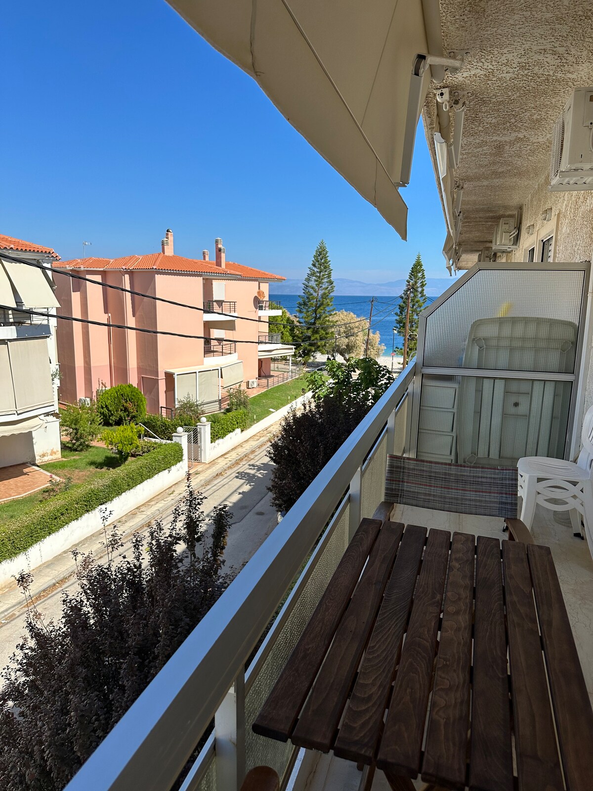A balcony view features a wooden table complemented by two white chairs overlooking a street lined with greenery. In the background, buildings and trees are visible, with the sea extending towards the horizon beneath a clear blue sky.