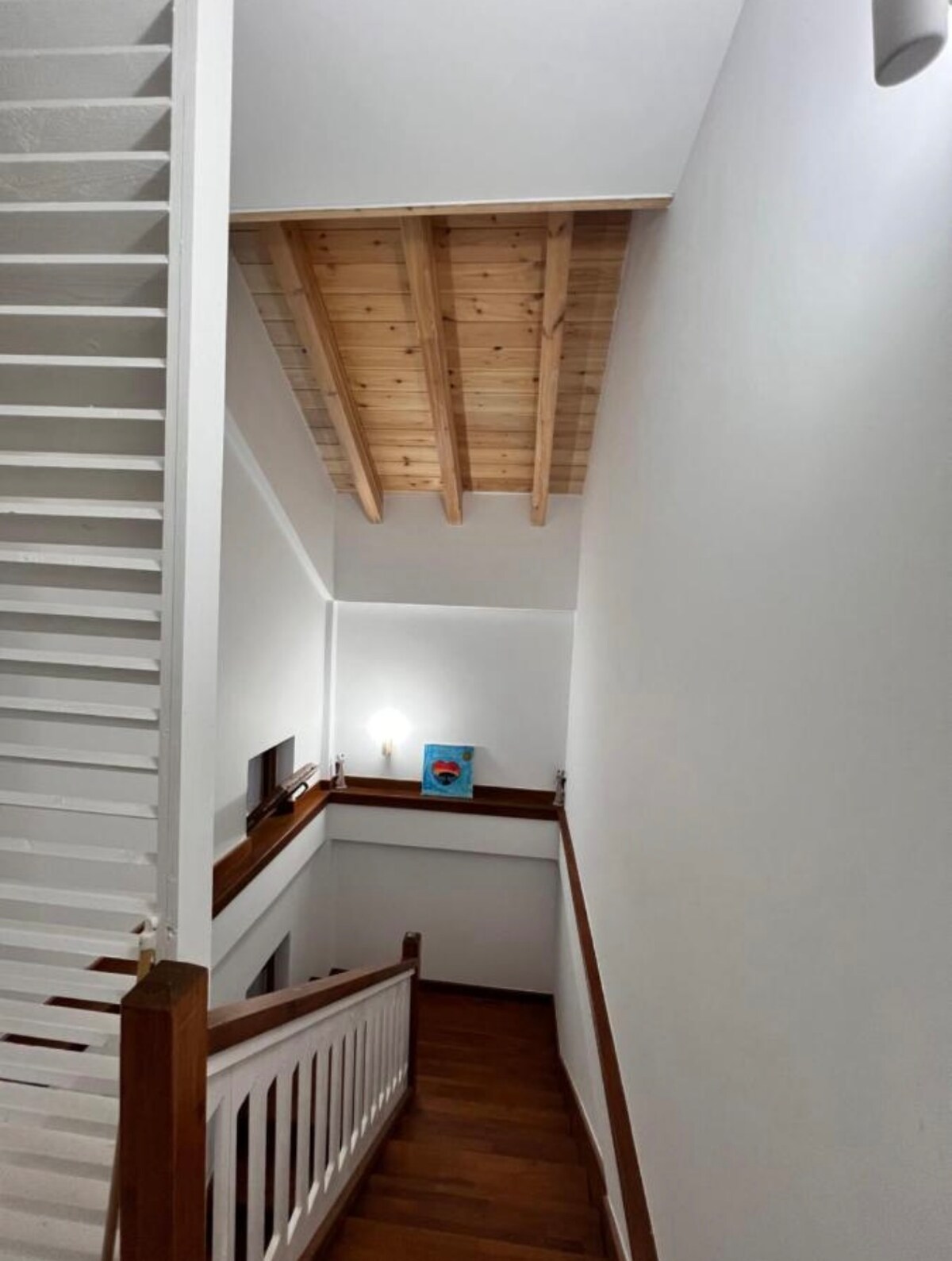 A staircase is featured with wooden railings and a polished floor. Light wood beams are visible on the ceiling, creating a spacious feel. The surroundings are painted in white, enhancing the brightness of the area. A small television is positioned on a ledge near the landing.