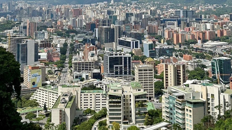 A panoramic view of an urban landscape showcasing a dense cluster of buildings, with varying architectural styles and heights. Green spaces are discernible among the structures, and distant mountains provide a natural backdrop to the bustling city scene.