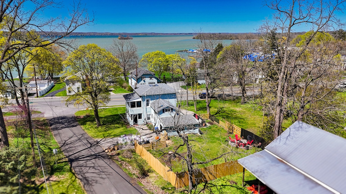 A sweeping view of the property reveals a spacious yard surrounded by trees and a newly fenced area. The lake gleams in the background, with boats visible at the marina. The neighborhood features well-maintained roads and nearby homes under a clear blue sky.