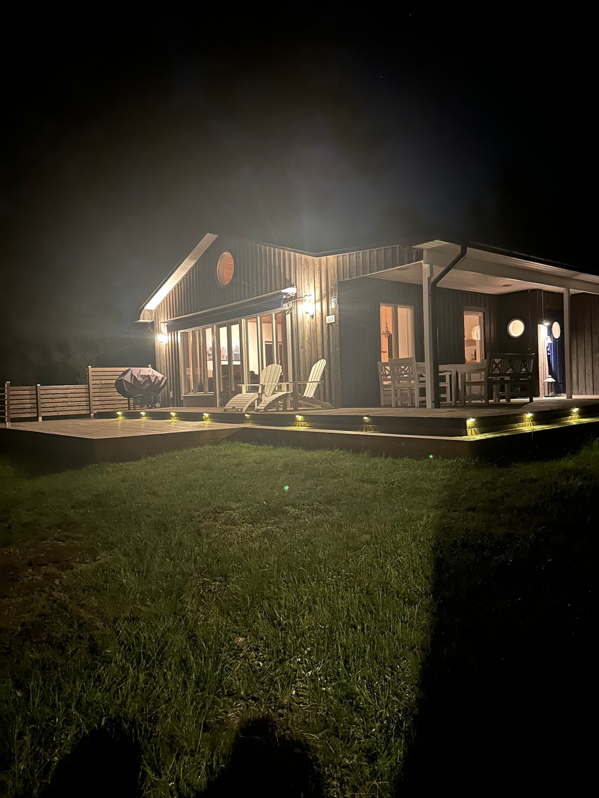 The cozy exterior of the house is illuminated at night, showcasing wooden siding and a welcoming deck. Two outdoor chairs are positioned on the deck, and soft lighting highlights the surrounding greenery. Large windows reflect light from within, creating a tranquil ambiance.