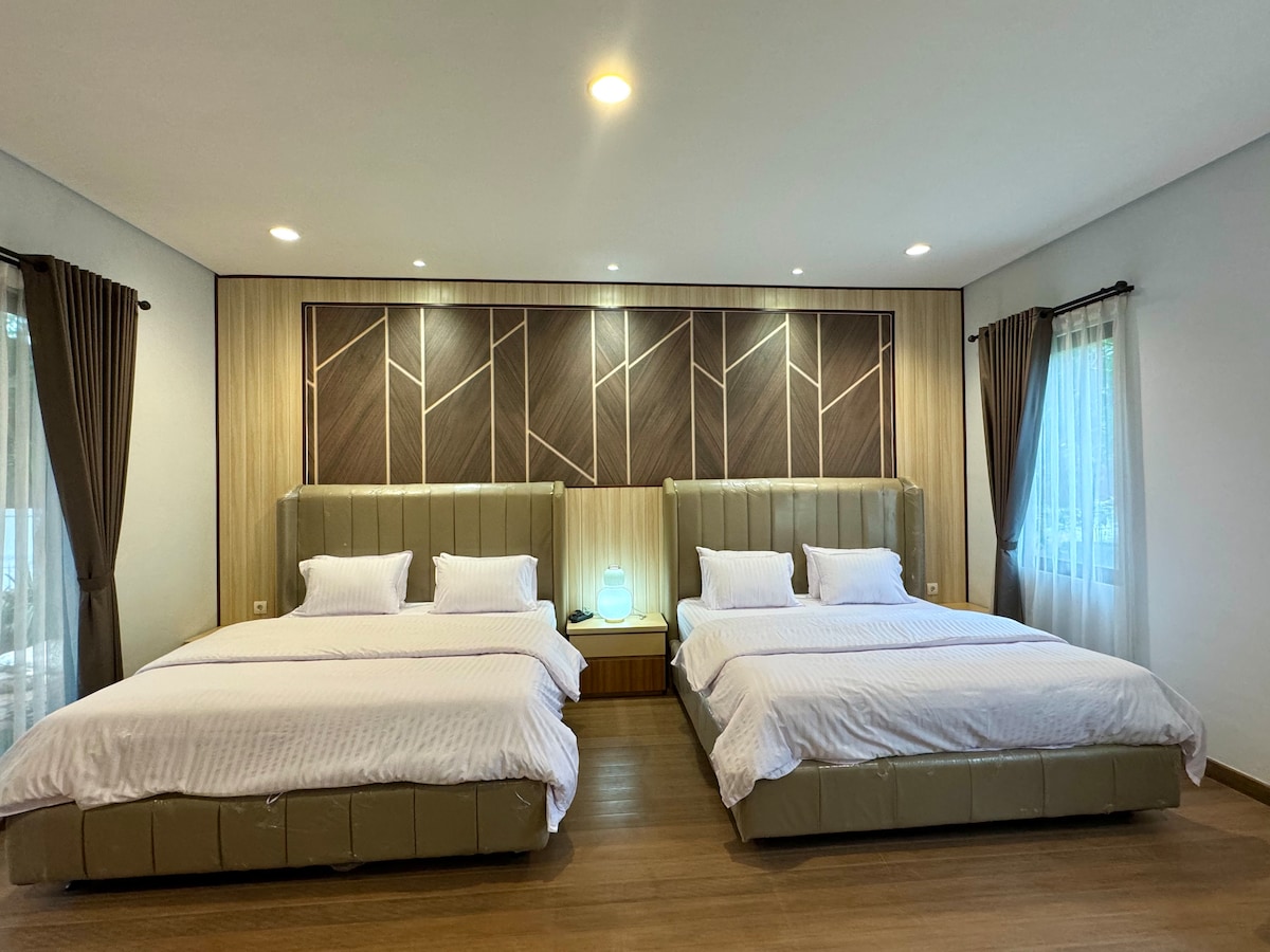 A spacious sleeping area is featured, showcasing two large beds with neutral-toned bedding. Soft ambient lighting is provided by bedside lamps, and natural light filters through curtains at the windows. The modern decor includes a patterned wooden accent wall, adding a touch of elegance.