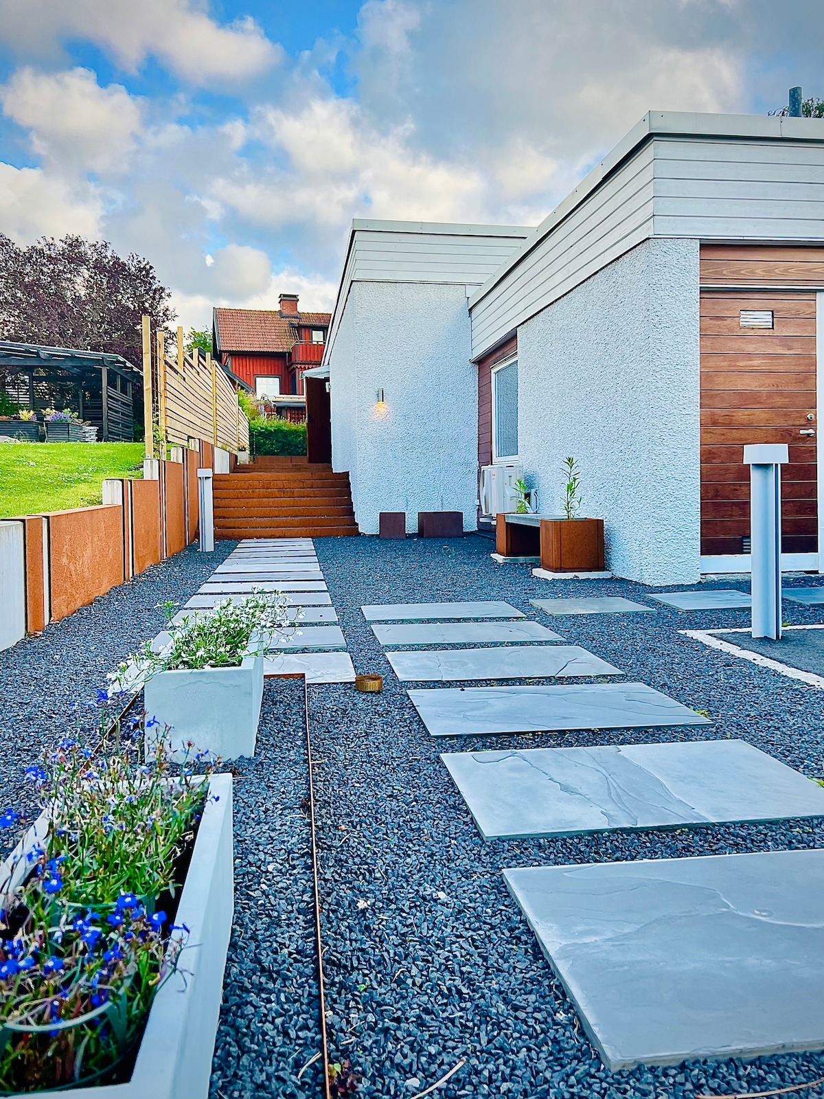 A neatly arranged pathway is lined with rectangular stone slabs, leading towards the villa. Flower beds featuring colorful plants are positioned along the sides. The exterior showcases a combination of white walls and wooden accents, set against a backdrop of greenery.