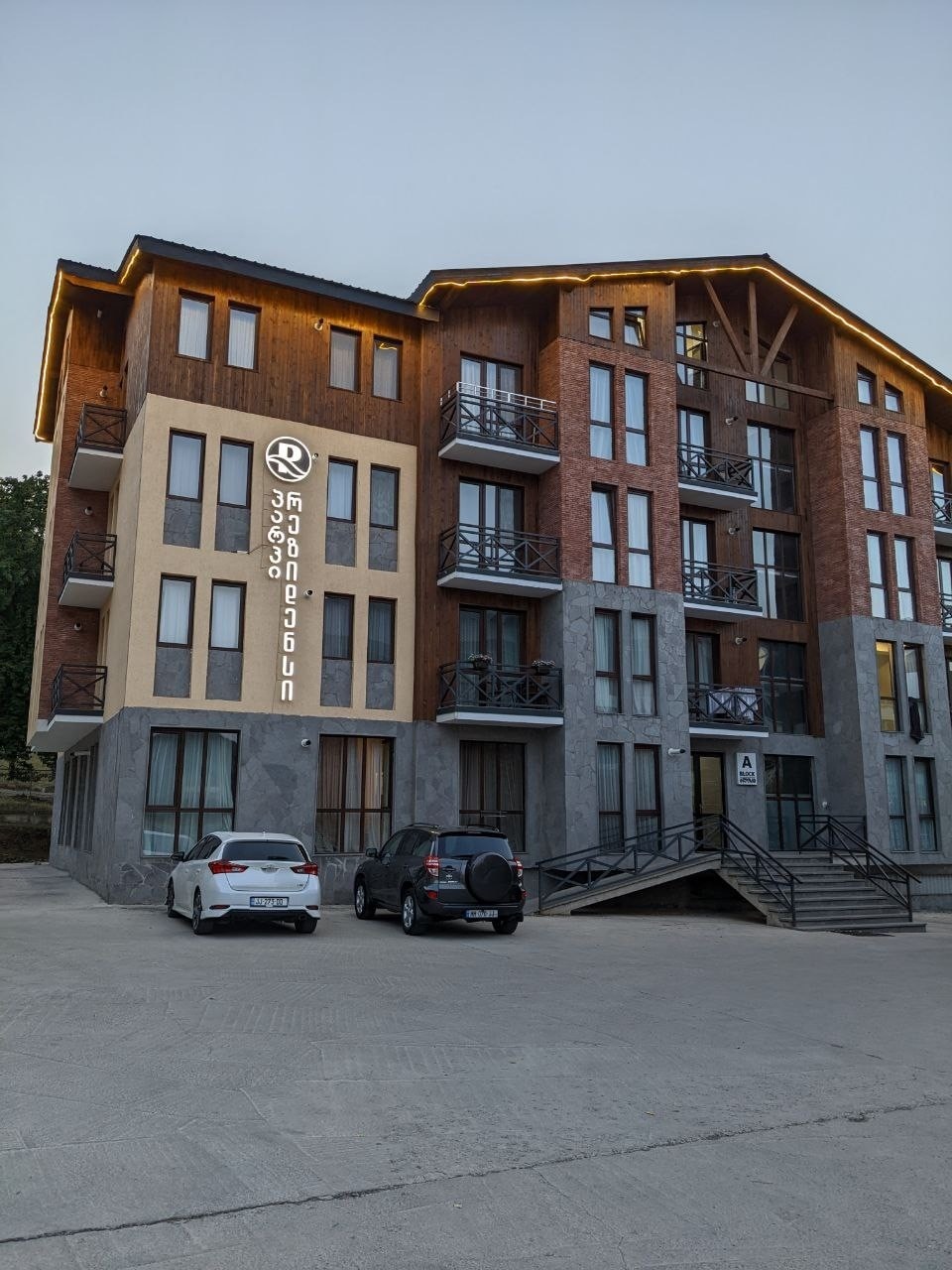 A five-story apartment complex is depicted, featuring a mix of wooden and stone façade. Large windows provide views of the surrounding area. The entrance is accessible by a staircase, with outdoor parking visible. The building's name is prominently displayed on the front.
