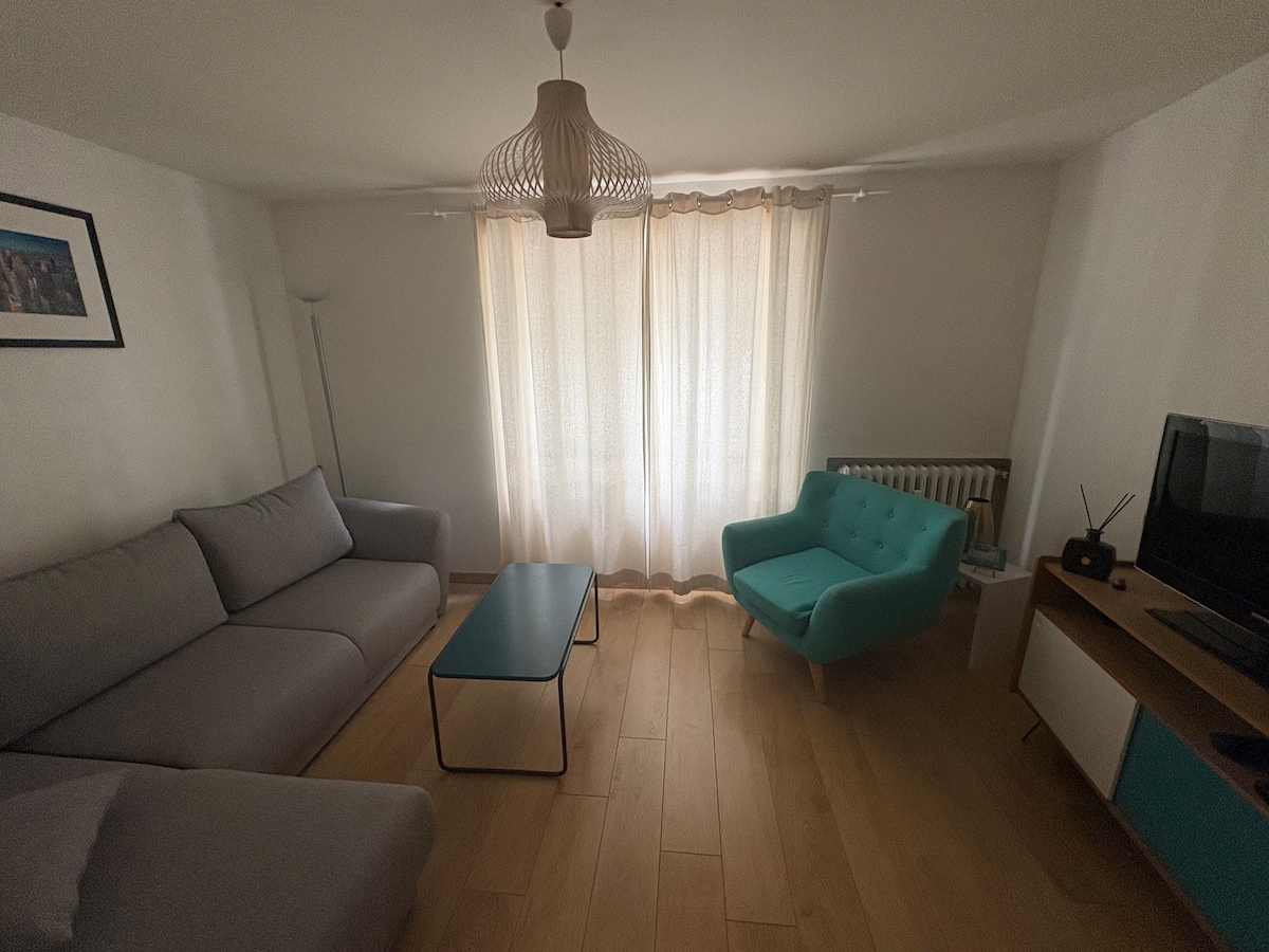 The living area is furnished with a light grey sofa and a turquoise armchair, accompanied by a modern coffee table. Natural light filters through the sheer curtains, illuminating the warm wooden flooring. A television sits on a minimalist media unit, enhancing the inviting and functional layout.