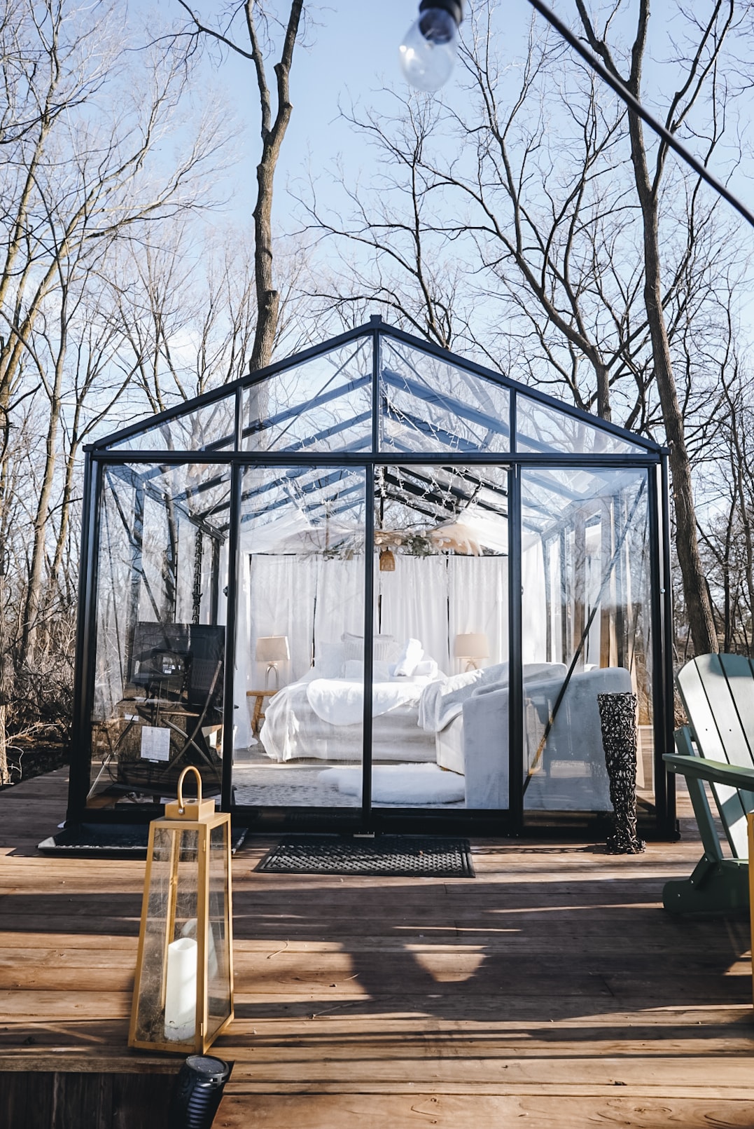 A glass house reflects natural surroundings, enclosed by trees and sunlight. The interior features a spacious bed draped in white linens, with cozy seating visible nearby. A wooden deck extends outwards, complemented by outdoor furniture and decorative lanterns.