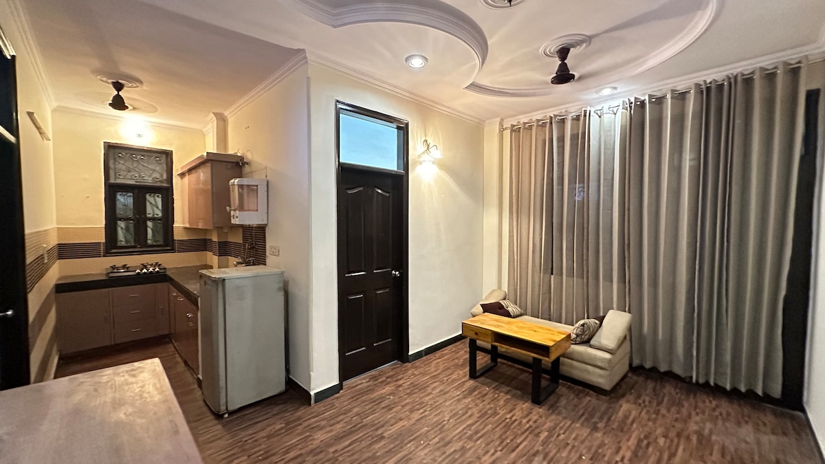 An open living space is seen, featuring a light wooden coffee table positioned in front of a soft seating area. Large windows are adorned with sheer curtains, allowing natural light to fill the room. A modern kitchen area is partially visible, showcasing appliances and cabinets.