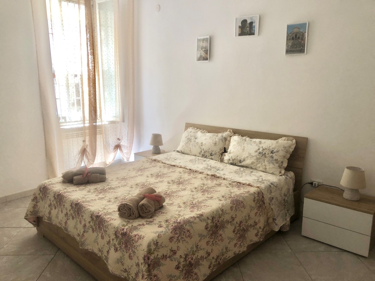 A serene bedroom features a spacious bed adorned with floral bedding. Soft lighting is provided by bedside lamps on either side. Wall-mounted photographs add a subtle decorative touch, while natural light filters through sheer curtains, creating a calm, inviting environment.