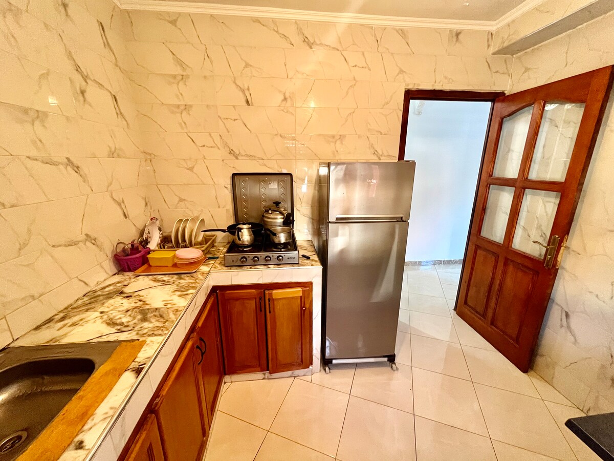 The kitchen features sleek marble walls and a functional layout. A stainless steel refrigerator is positioned beside a stovetop with pots visible on the burners. Wooden cabinets provide storage space, and a sink is integrated into the countertop, enhancing the kitchen's utility.