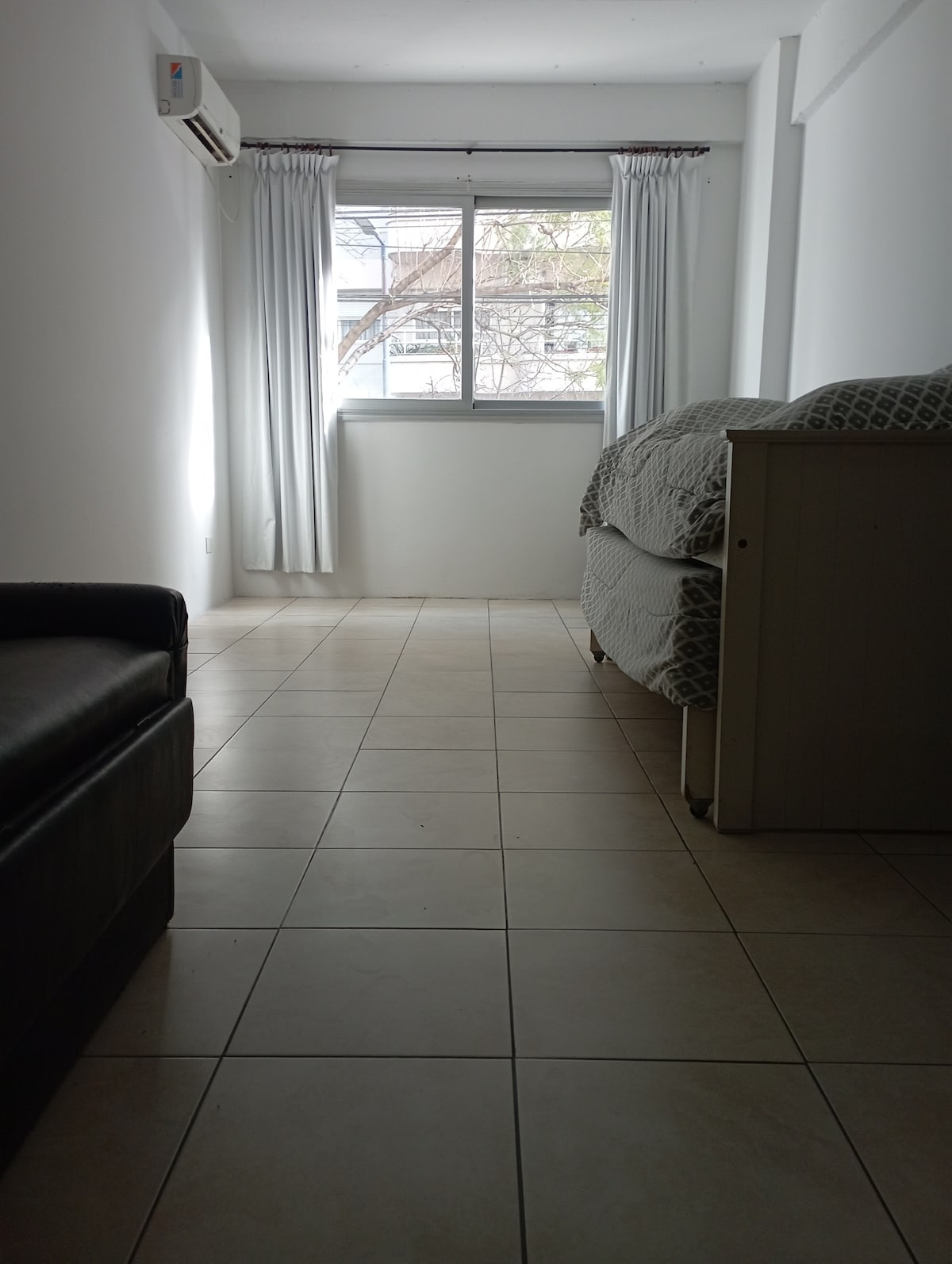 A well-lit interior space features a large window with light curtains allowing natural light to enter. A neutral-colored couch is positioned to the left, while a cozy sleeping area is visible to the right, showcasing a neatly made bed with a patterned coverlet.