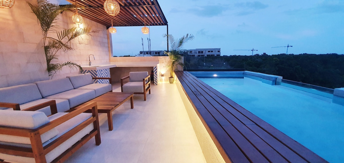 A rooftop area is equipped with light-toned seating and a wooden table, offering a relaxed atmosphere. A rectangular pool reflects the evening sky, while soft lighting enhances the space. Lush greenery is subtly incorporated, adding a touch of nature to the contemporary design.