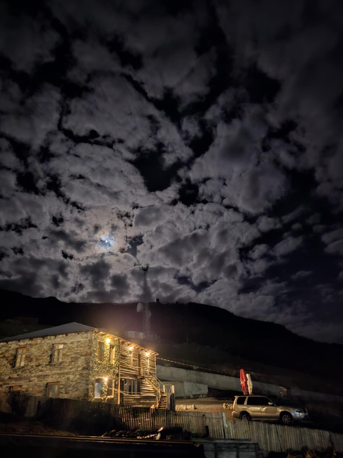 A stone guesthouse is illuminated by soft warm light, positioned against a dramatic night sky filled with billowing clouds. The moon is visible, casting a gentle glow over the surrounding mountains, enhancing the tranquil atmosphere of the setting.