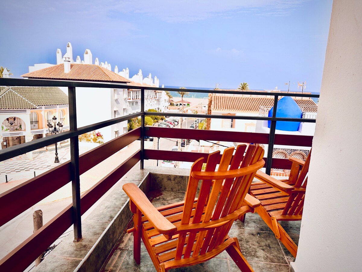 A private patio features two wooden chairs positioned to overlook the nearby street and ocean. The railing provides a clear view of the landscape, which includes white buildings and palm trees under a bright sky.
