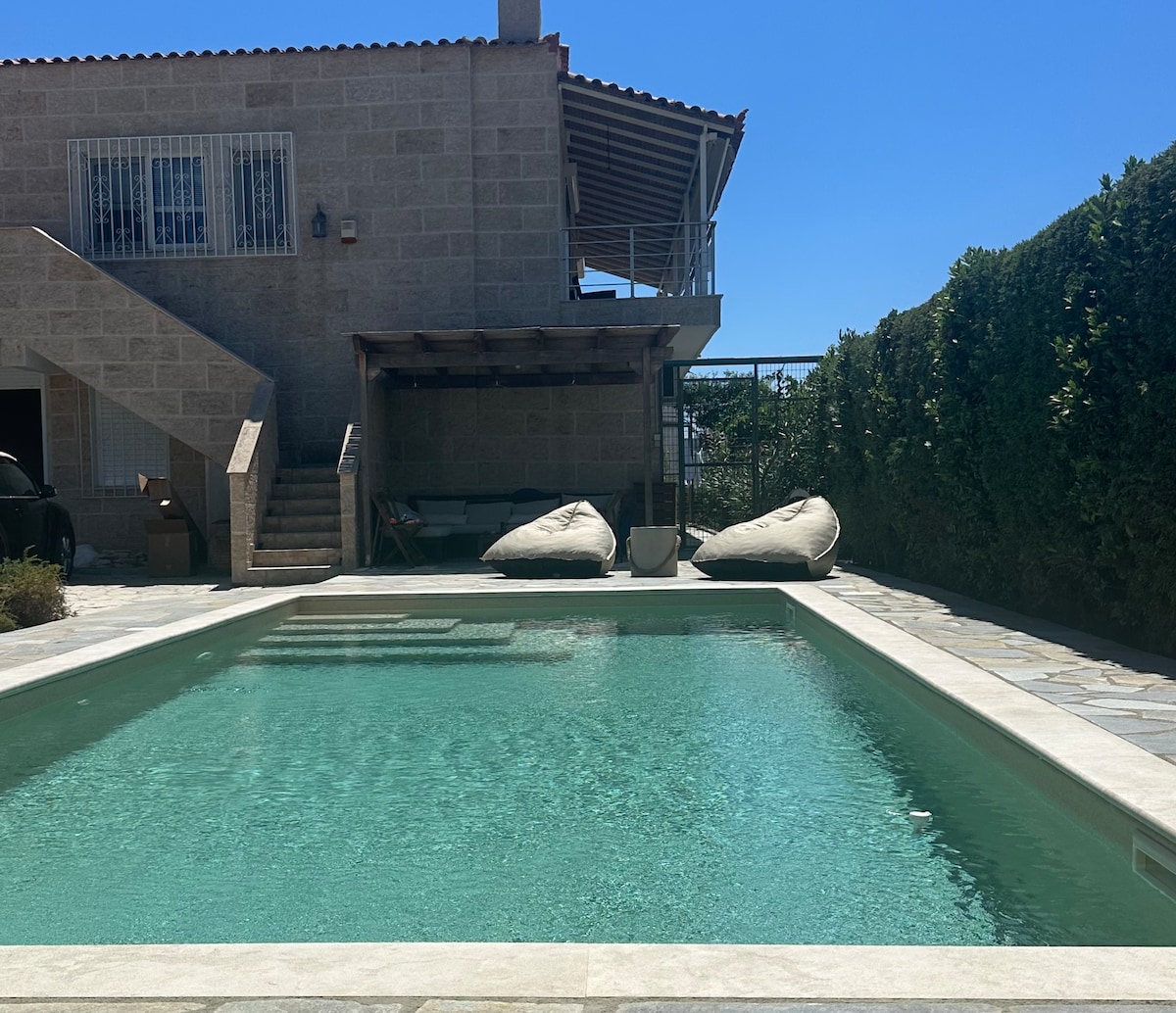A private saltwater pool is surrounded by a spacious patio area, featuring two bean bag chairs. The house's exterior is visible in the background, with a staircase leading to an upper level. Lush greenery provides a natural border, enhancing the serene outdoor space.