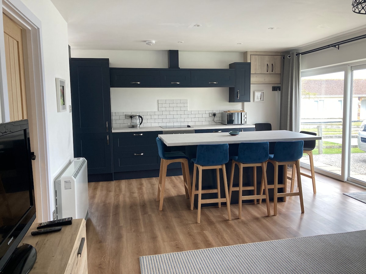 The open plan kitchen and dining area features modern cabinetry in deep blue tones. A central island provides seating with blue upholstered stools. Light streams through patio doors, enhancing the airy feel. Wood flooring extends throughout the space, complemented by a television in the corner.